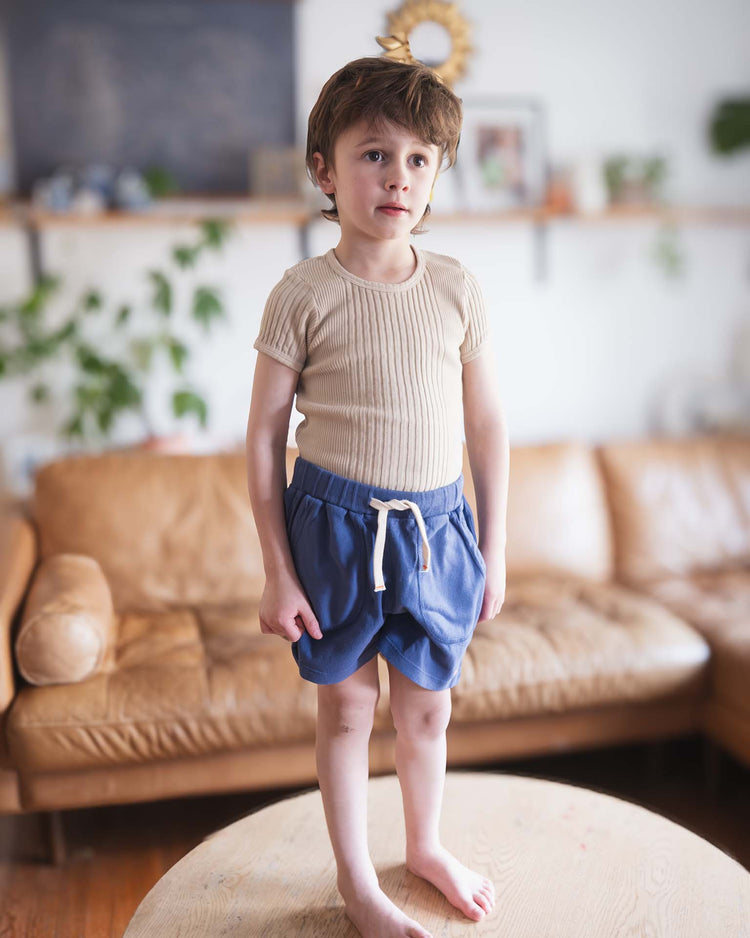 A young child with light brown hair stands barefoot on a wooden table, wearing a beige ribbed shirt and Sunchild's merida shorts in obscur, featuring a dip-dyed look, drawstring waist, and cotton fabric. A tan sofa and blurred plants are in the background.