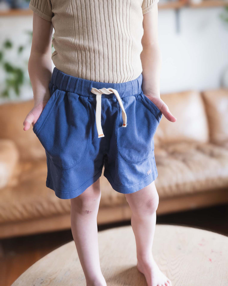 A child stands barefoot on a round wooden table, wearing a beige ribbed shirt and Sunchild’s Merida shorts in Obscur, hands in pockets. A tan couch and plants are visible in the blurred background.