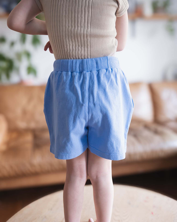 A young Sunchild wearing Merida shorts in Mare by Sunchild stands barefoot on a wooden table, with a brown sofa and green plants blurred in the background.
