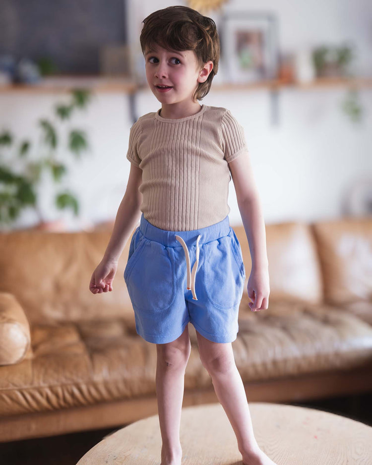 A young child with short brown hair stands barefoot on a wooden table, wearing the loutro tee in craie by sunchild and blue shorts. A brown leather sofa and plants can be seen in the softly focused background.
