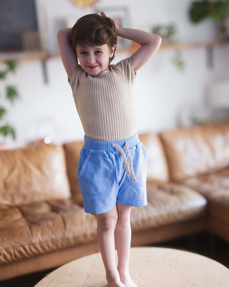 A young Sunchild with short brown hair stands barefoot on a wooden table, smiling with arms raised behind their head, wearing a beige shirt and sunchild merida shorts in mare. A brown sofa and plants appear in the background.