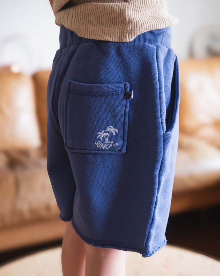 A person wearing sunchild pico shorts in obscur—blue fleece cotton with a drawstring waist and front pocket featuring a palm tree and YNCAL text—stands indoors with a tan couch in the background.