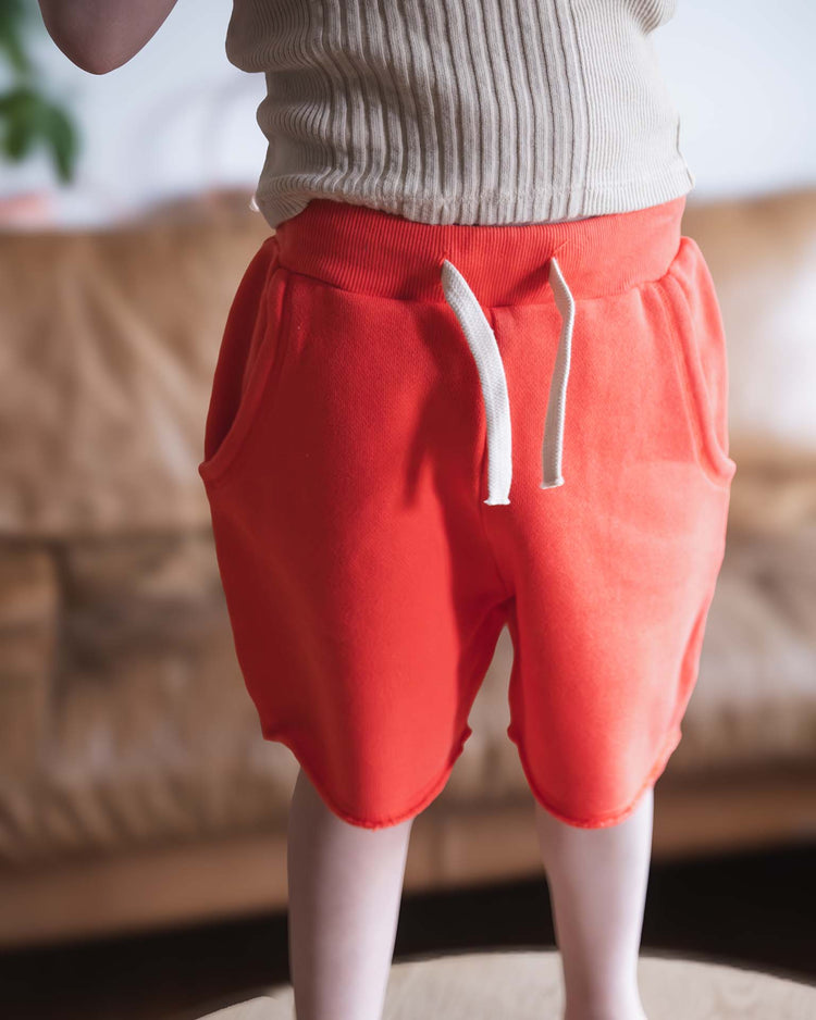 A Sunchild is indoors wearing pico shorts in tangerine by sunchild, featuring a bright orange color and drawstring waist, paired with a ribbed beige top and hands out of frame.