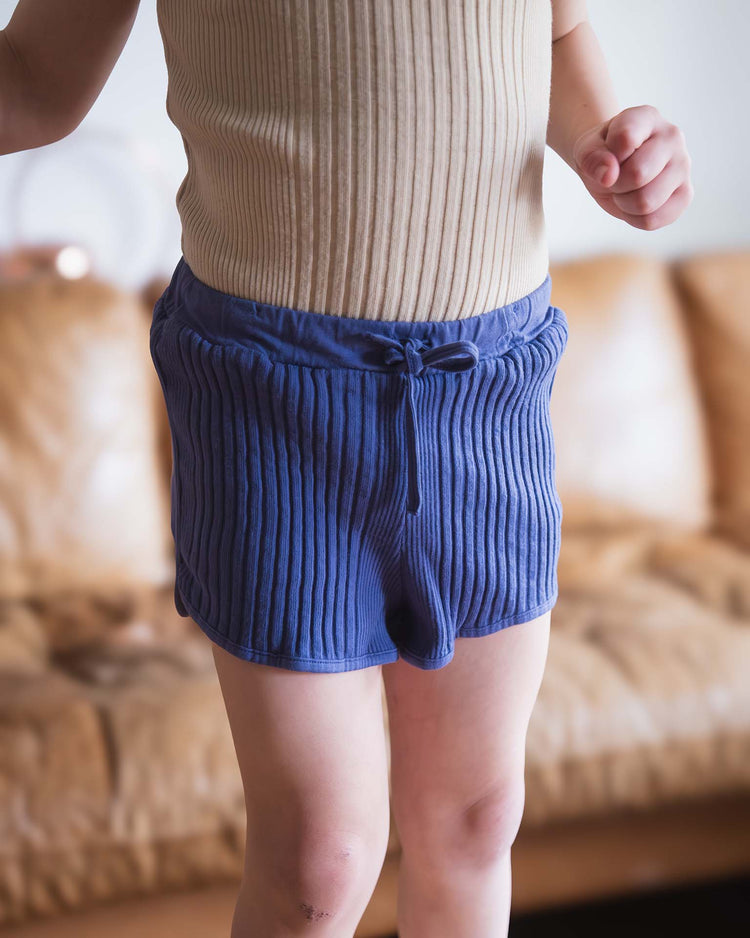 A child stands indoors in front of a tan couch, wearing a beige ribbed tank top and Sunchild rio shorts in obscur with a comfy drawstring waist. Only the child’s torso and legs are visible.