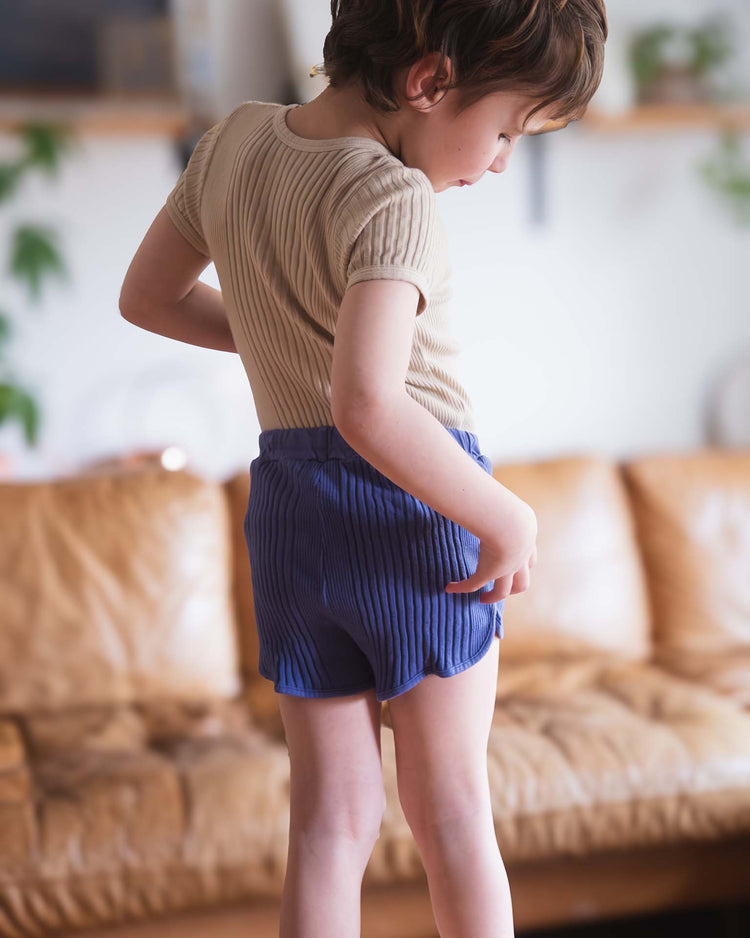 A young child with short brown hair, wearing a beige ribbed top and Sunchild's rio shorts in obscur with a drawstring waist, stands sideways on a tan leather couch and looks down at their hand against a blurred background.