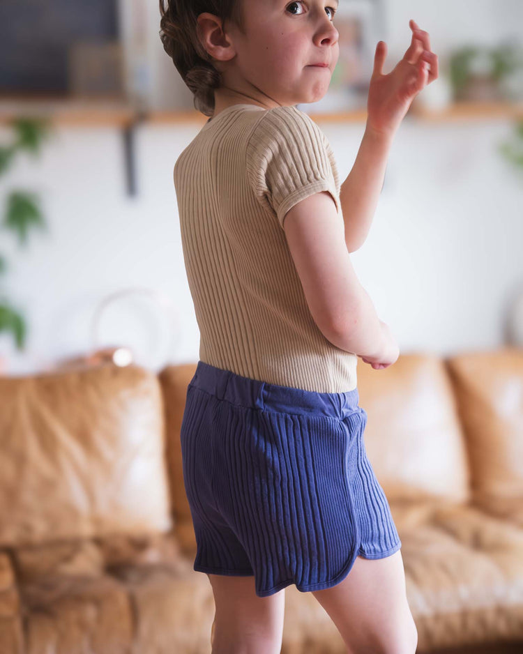 A young child wears a beige ribbed shirt and Sunchild rio shorts in Obscur with a drawstring waist, standing indoors before a brown sofa and glancing over their shoulder with one hand raised.