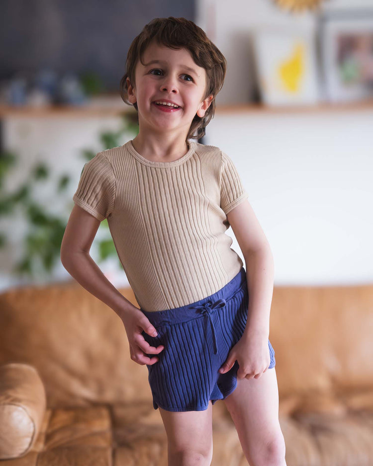 A young child with short brown hair smiles while standing on a brown leather couch, wearing a beige ribbed shirt and sunchild rio shorts in obscur with a drawstring waist. Softly blurred plants and wall art appear in the background.