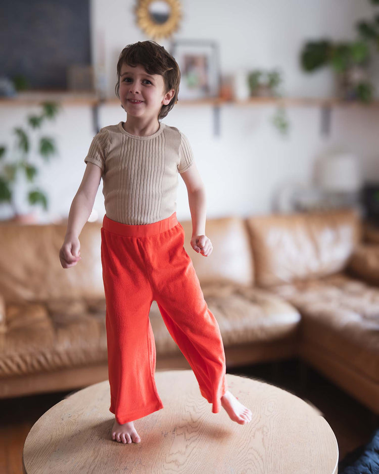 A young child with short brown hair, wearing a beige shirt and Sunchild bluff pants in tangerine, stands barefoot on a round wooden table in a cozy living room with a brown sofa and green plants in the background.