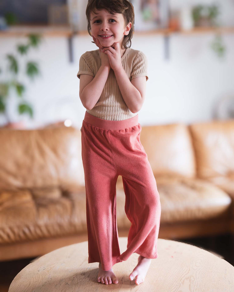 A young child, barefoot on a wooden table, smiles with hands to chin. They wear a beige shirt and Sunchild bluff pants in cacao, with a brown couch and green plants softly blurred in the background.
