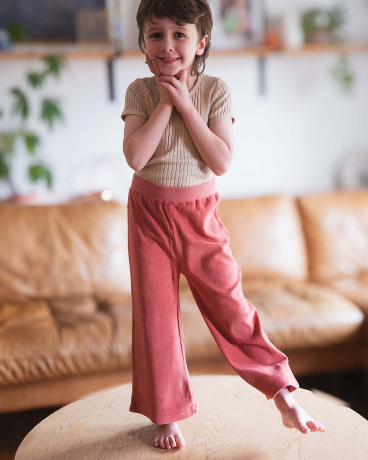 A young child stands barefoot on a wooden table, smiling shyly with hands under their chin, wearing a beige ribbed shirt and sunchild bluff pants in cacao. A brown sofa and plants are visible in the background.