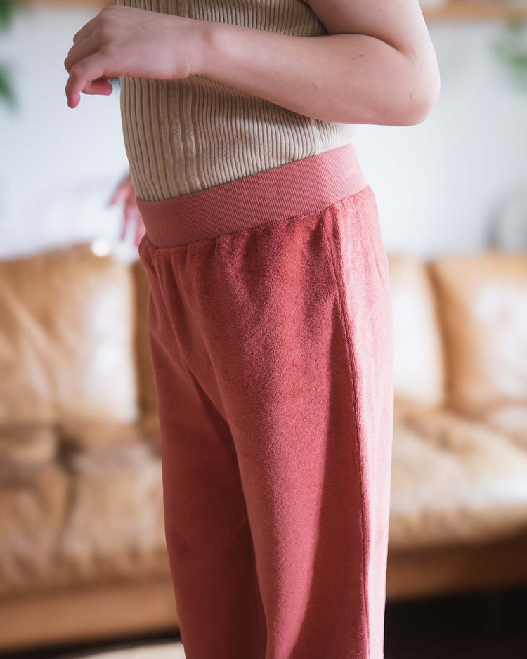 A person wears Sunchild's bluff pants in cacao and a beige ribbed top, standing indoors with a tan sofa behind them; only their torso and arms are visible.