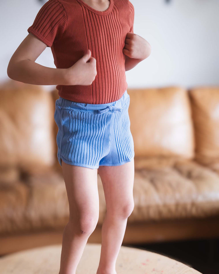 A child stands on a wooden surface in a rust ribbed shirt and Sunchild’s rio shorts in mare—light blue stripes with a drawstring waist. A tan leather couch is blurred in the background, and the child’s face is not shown.