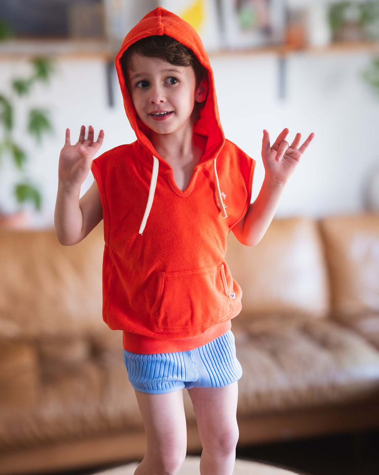 A young child smiles indoors with hands raised, wearing a Sunchild sitges sleeveless sweatshirt in tangerine and blue striped shorts. A tan couch and green plants appear in the blurred background.