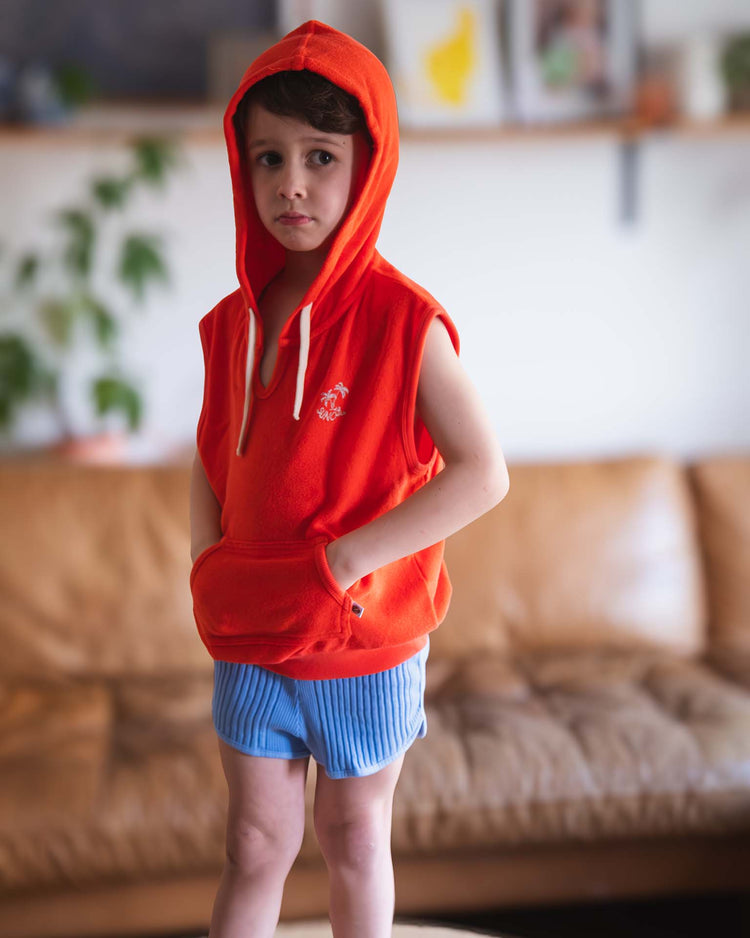A young child wears a Sunchild sitges sleeveless sweatshirt in tangerine and blue shorts, standing indoors before a brown sofa with hands in the hoodie pocket. A plant and shelves appear blurred in the background.