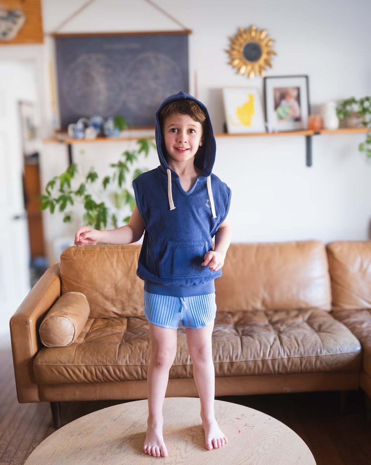 A young child in the sunchild sitges sleeveless sweatshirt in obscur stands barefoot on a round wooden table in a living room with a tan leather couch and wall decorations in the background.