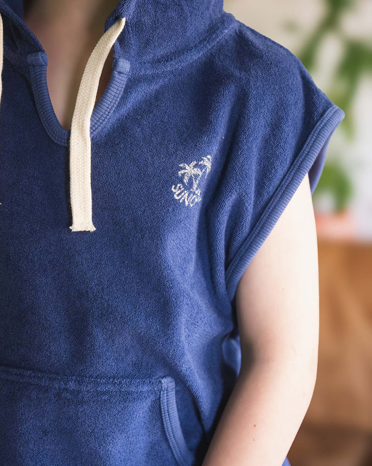 A person wears the sitges sleeveless sweatshirt in obscur by sunchild, a blue dip-dyed hoodie with a beige drawstring, white palm tree graphic, and SYNC embroidered on the chest; background is softly blurred.