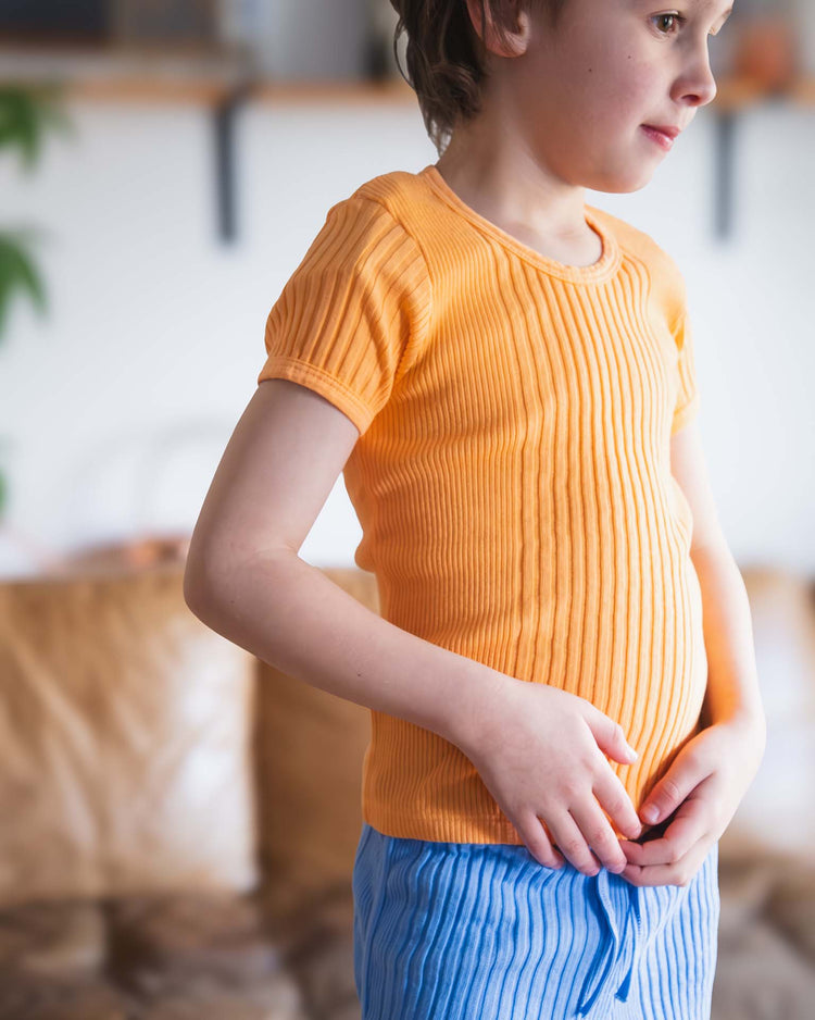 A child stands indoors, looking to the side with hands clasped, wearing the Sunchild loutro tee in inca and orange and blue striped pants. The softly blurred background adds a gentle touch.