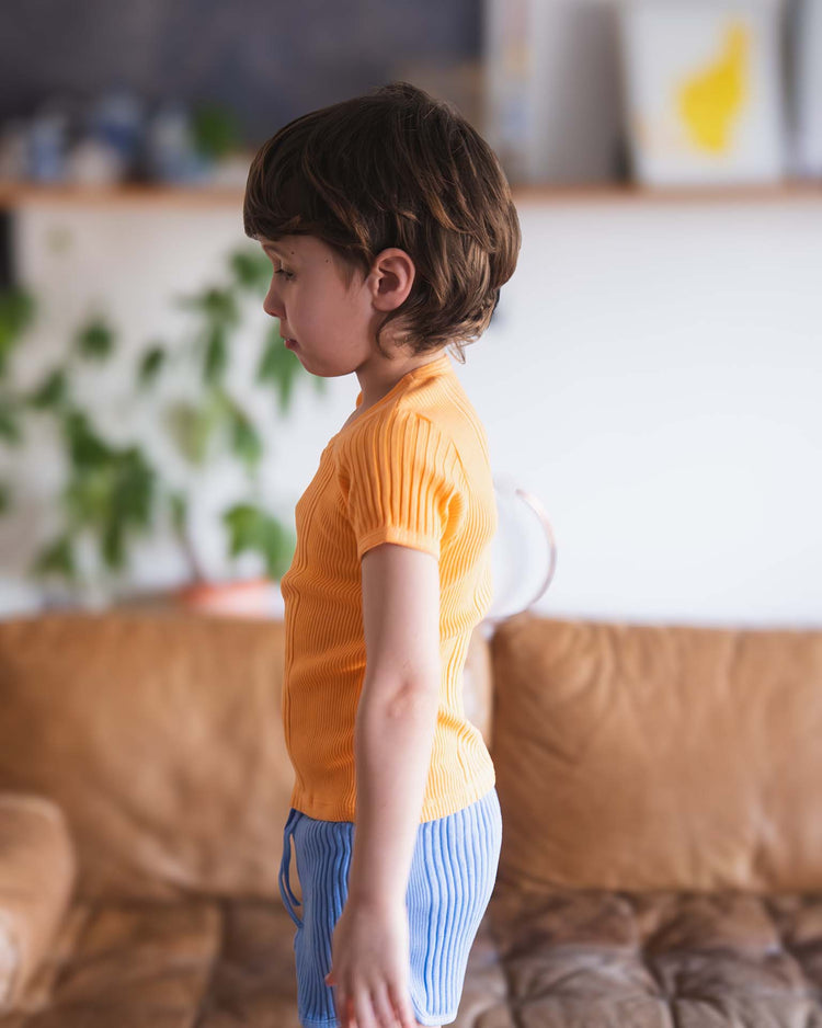 A young child with brown hair stands in profile on a brown couch, wearing the sunchild loutro tee in inca and blue striped shorts. A blurred plant and shelves with books and decor appear in the background.