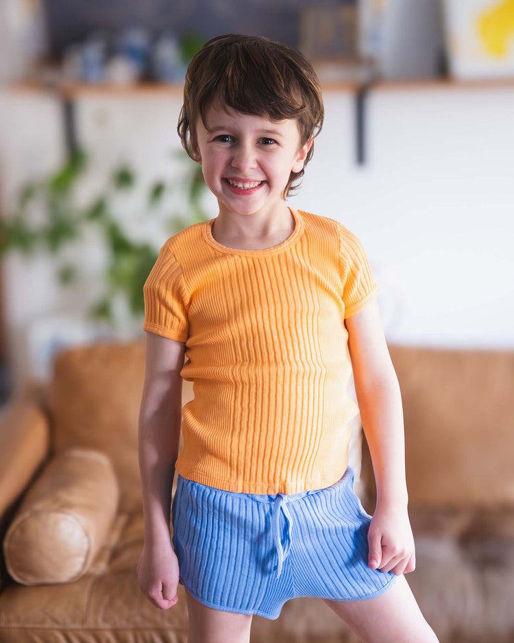 A smiling young child with short brown hair wears a sunchild loutro tee in inca and blue ribbed shorts, standing indoors before a brown sofa with green plants in the background.