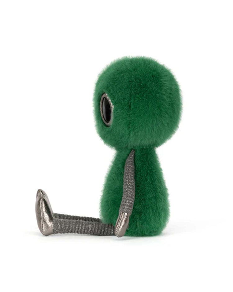 A jellycat zyllan alien plush toy with a fuzzy green head and body, large black eyes, and silver metallic limbs sits upright facing left against a white background.