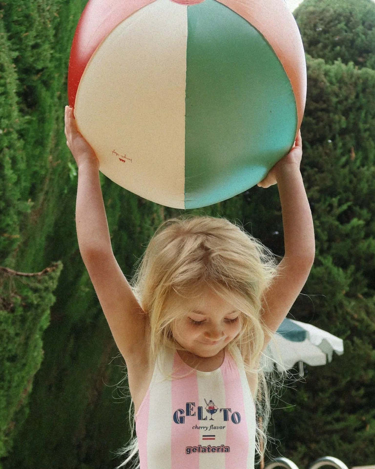 A young girl with long blonde hair holds a large konges sløjd beach ball in multi above her head while wearing a striped swimsuit, with lush green trees in the background.