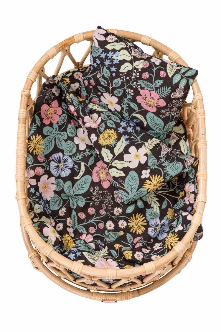 The Poppie Crib Botanical Collection by poppie toys is a woven basket featuring a black fabric adorned with a floral pattern of various colorful flowers and leaves, handcrafted by skilled craftsmen.