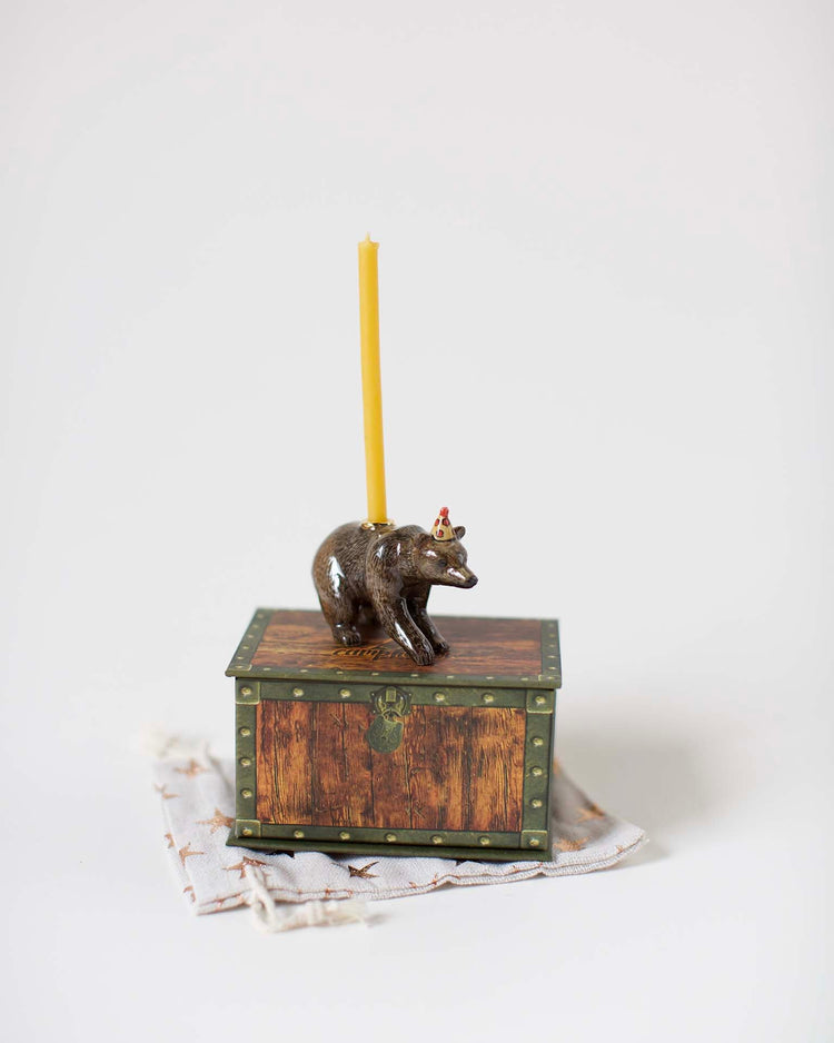A decorative hand-painted candle holder shaped like a brown bear on top of a small, ornate wooden box, featuring a green floral design, with a lit yellow candle.
