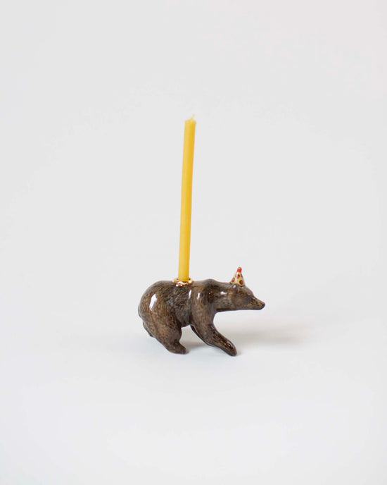 A small metal figurine of a camp hollow brown bear cake topper with a bright yellow candle protruding from its back, set against a plain white background.