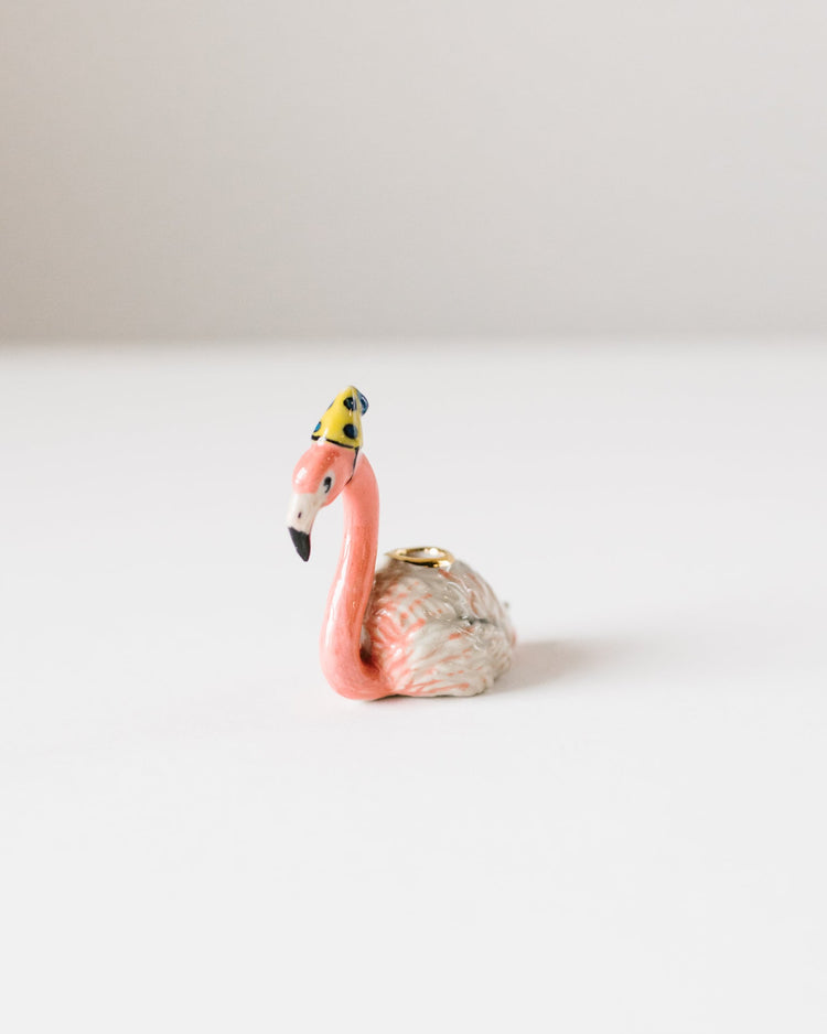 A small ceramic Camp Hollow flamingo cake topper, featuring a pink body and a tiny yellow hat, positioned against a plain white background.