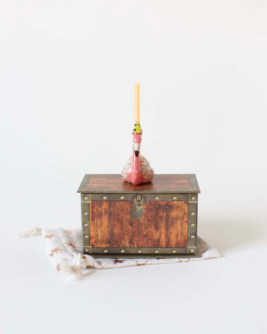 A small, antique wooden chest with metal accents and a single flamingo cake topper mounted on a ceramic candle holder, placed on a light background. (Brand Name: camp hollow)