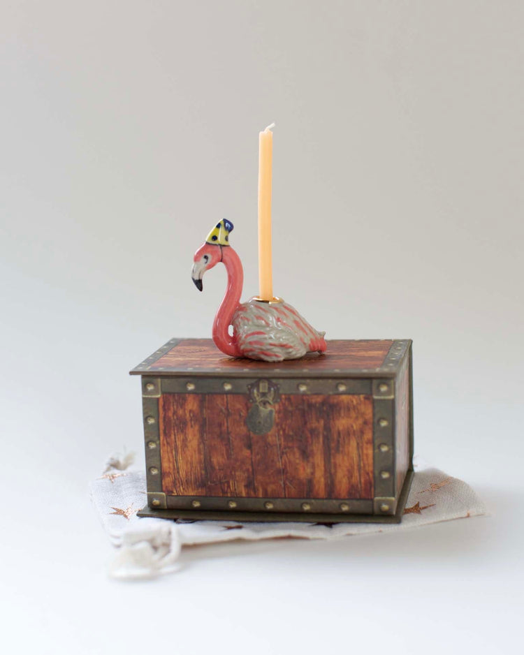 A whimsical ceramic Camp Hollow flamingo cake topper candle holder with a tall yellow candle, positioned on a small wooden treasure chest on a light background.