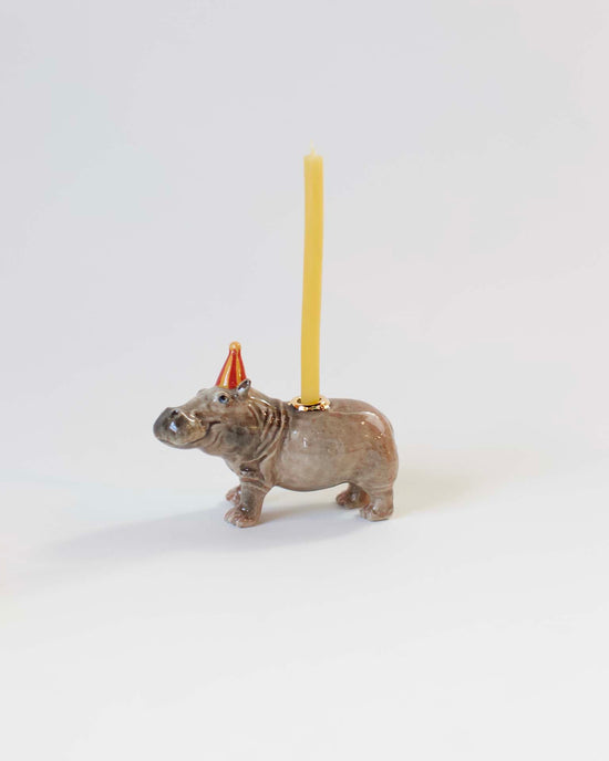 A hand-painted, ceramic camp hollow hippo cake topper with a yellow candle positioned on its back, set against a plain white background.