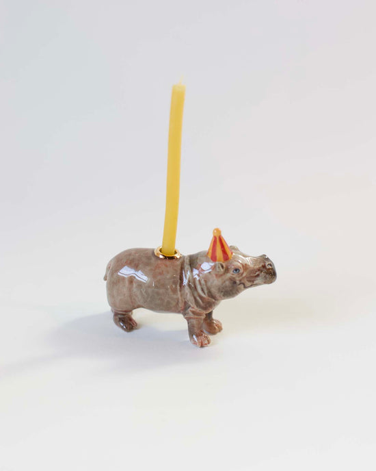 Camp Hollow hippo cake topper with a yellow candle mounted on its back, placed against a plain white background.