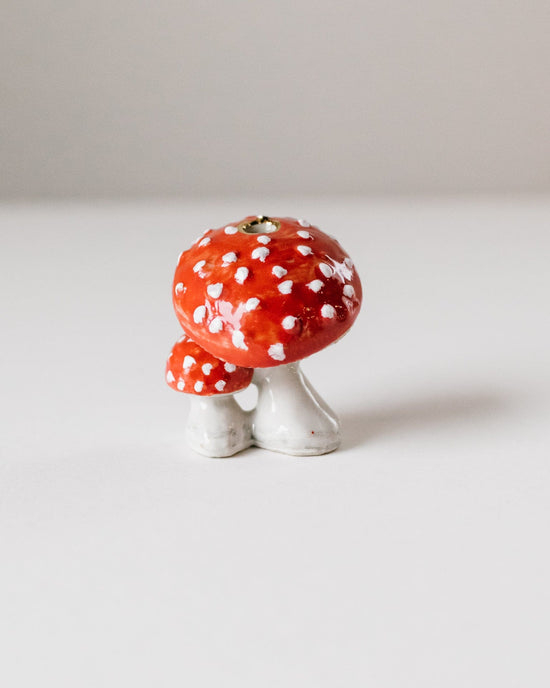 A hand-painted ceramic figurine of a red and white spotted mushroom cake topper with two white stems, displayed on a minimalistic grey background by Camp Hollow.