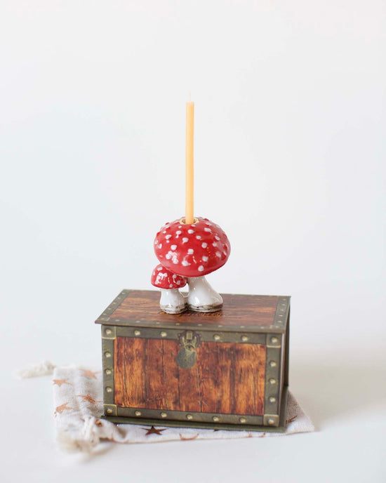 A hand-painted ceramic mushroom cake topper with a red and white cap, featuring a single orange candle on top, placed on a small wooden chest against a white background by camp hollow.