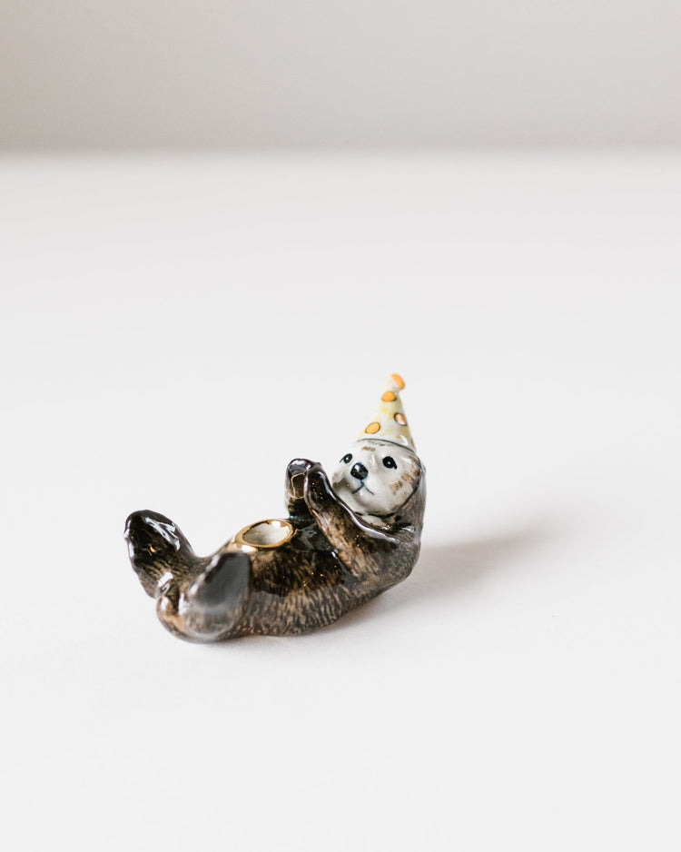 A small ceramic otter cake topper of a playful seal wearing a party hat, lying on its back against a white background.