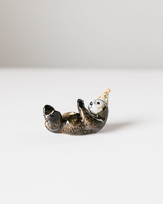 A ceramic otter cake topper of a sea otter lying on its back, wearing a small, colorful party hat, against a plain white background by Camp Hollow.