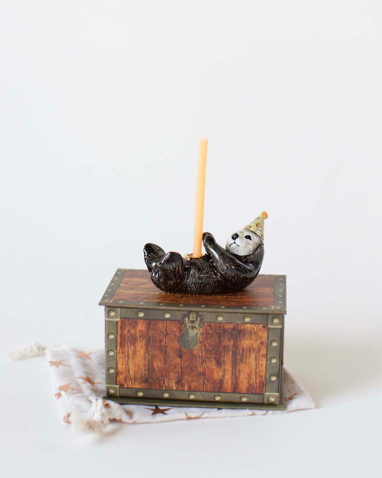 A ceramic otter cake topper, demonstrating heirloom-quality craftsmanship, wearing a party hat reclines on a small, ornate wooden chest, holding a beige candle upright.
