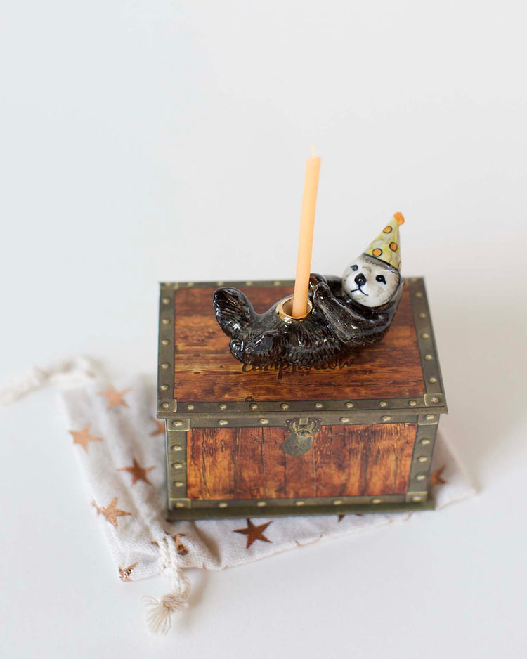 A camp hollow otter cake topper in a party hat holding a candle, atop a small wooden chest with star patterns, against a white background, showcases heirloom-quality craftsmanship.