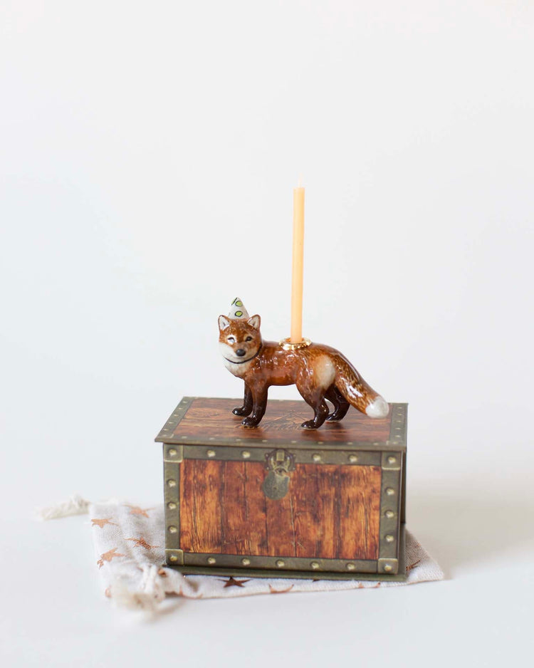 A hand-painted ceramic red fox cake topper from Camp Hollow on a small wooden chest, holding a pencil on its back, against a plain white background.