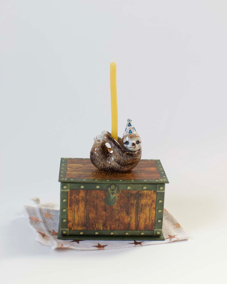 A small, whimsical camp hollow sloth cake topper wearing a party hat and holding a yellow candle, placed atop an ornate wooden box on a light background.