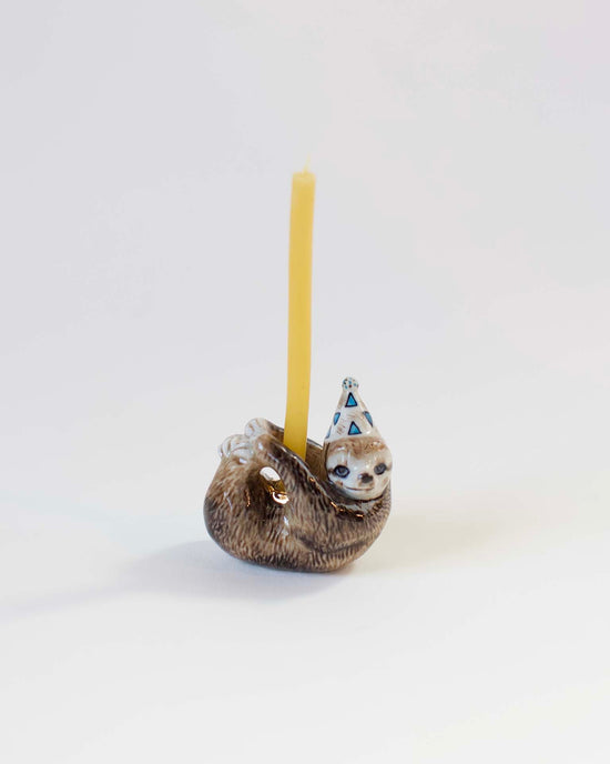A hand-painted ceramic Camp Hollow sloth cake topper wearing a party hat, holding a yellow candle, set against a plain white background.