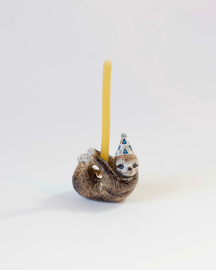 A hand-painted ceramic Camp Hollow sloth cake topper wearing a party hat, holding a yellow candle, set against a plain white background.
