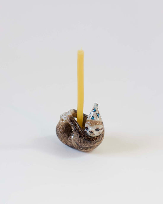 A hand-painted ceramic sloth-shaped candle holder with a yellow candle and a decorative blue and white party hat on a white background.