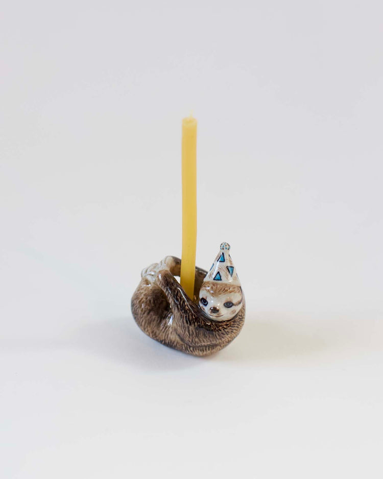 A hand-painted ceramic sloth-shaped candle holder with a yellow candle and a decorative blue and white party hat on a white background.