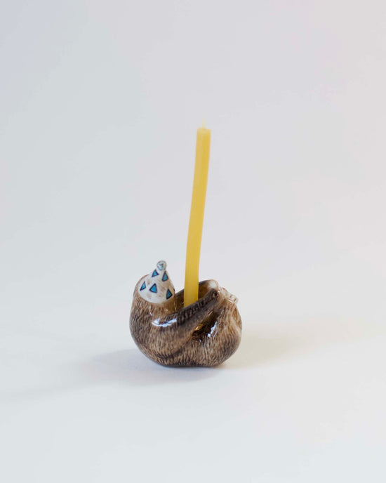 A yellow candle stands upright in a small, brown, ceramic sloth-shaped holder that is hand-painted with blue and white patterns, set against a plain white background. (Camp Hollow)