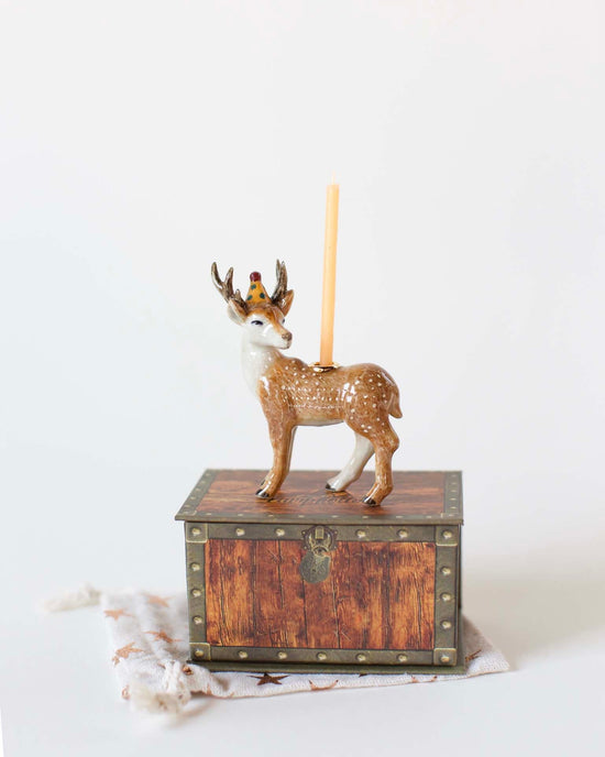 A Camp Hollow stag cake topper with a crown stands atop a small wooden chest, holding a single upright orange hand-painted candle holder.