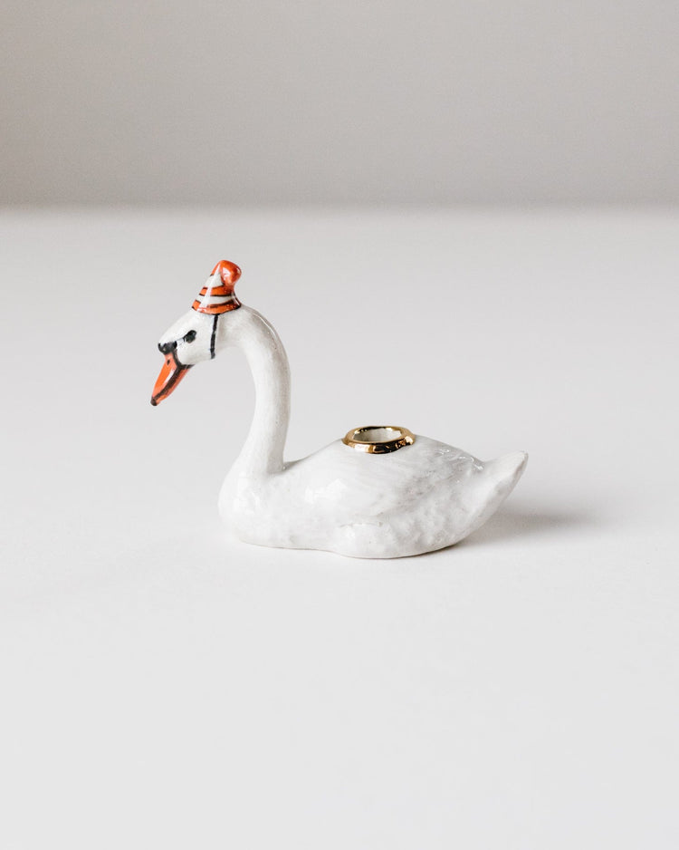 A Camp Hollow hand-painted porcelain swan cake topper wearing a small party hat, with a wedding ring placed on its back.