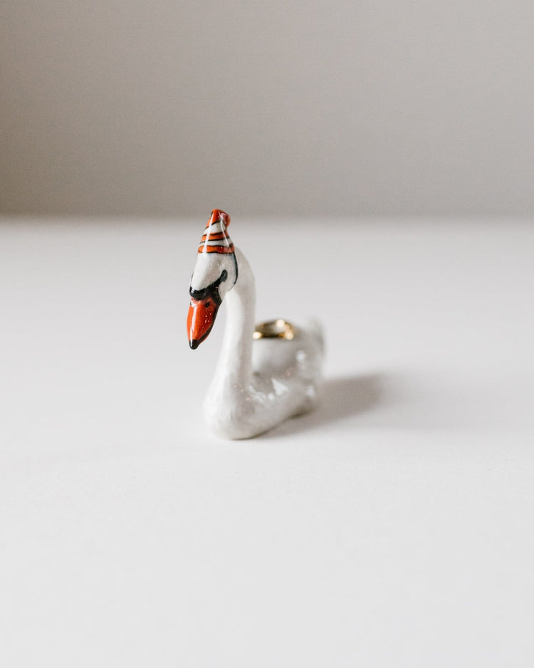 A small hand-painted ceramic figurine of a swan cake topper with a detailed face and golden accents, positioned on a plain white surface by Camp Hollow.