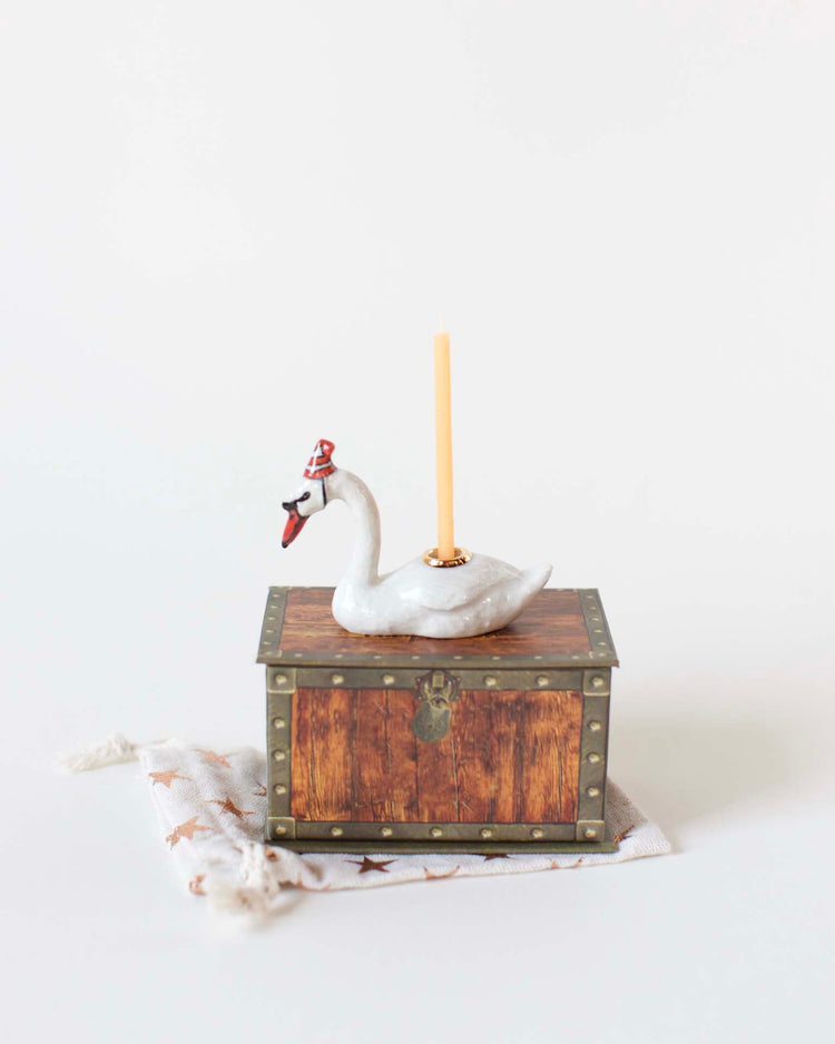 A hand-painted porcelain swan cake topper from Camp Hollow, with a pencil on a small wooden chest, placed on a light background.